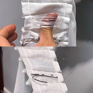 Close-up of white curtain pleating tape being pulled taut by a thumb, showing the fabric and hooks.