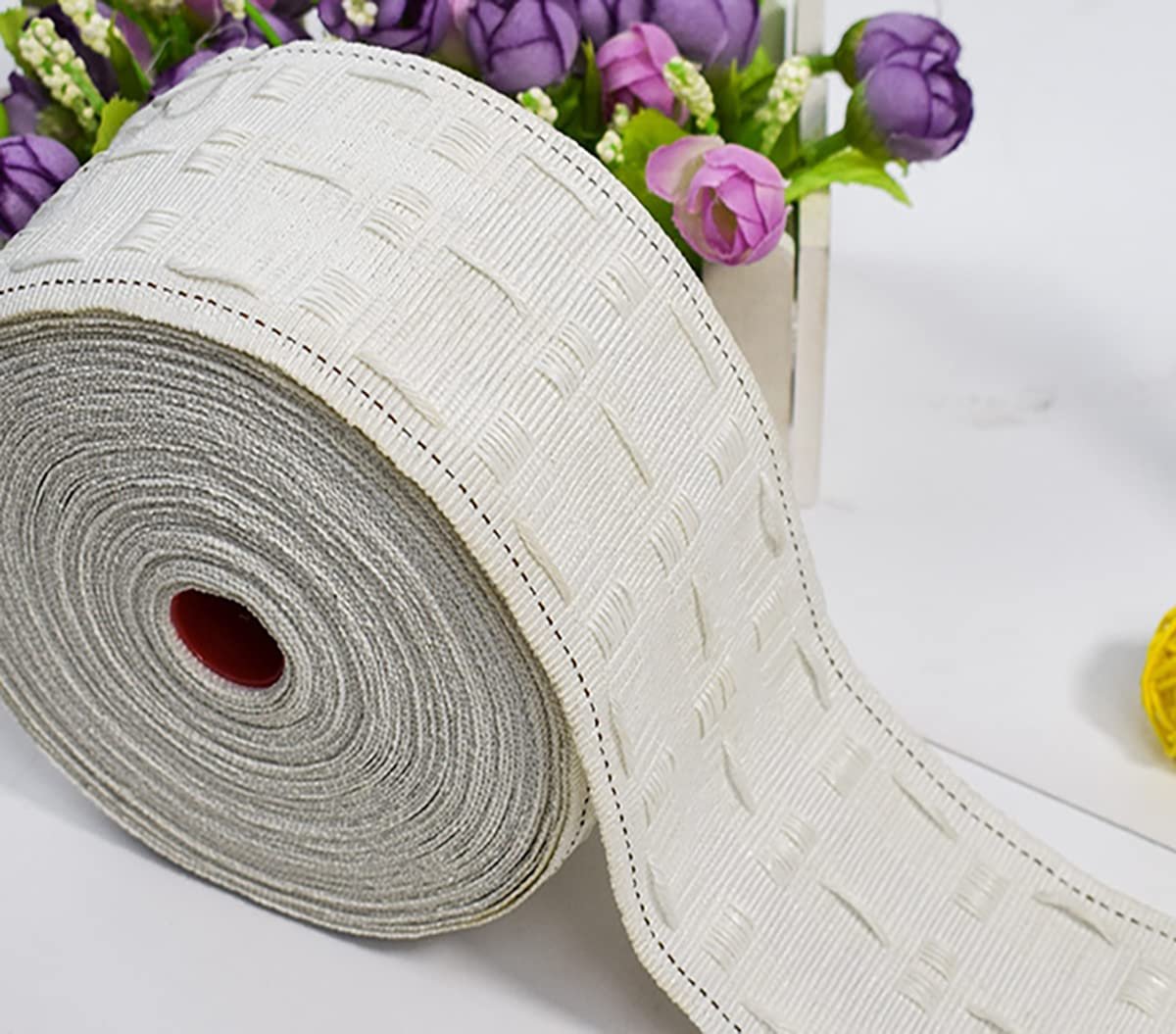Roll of white curtain pleating tape with a woven texture and stitched details, set against a white surface with decorative flowers.