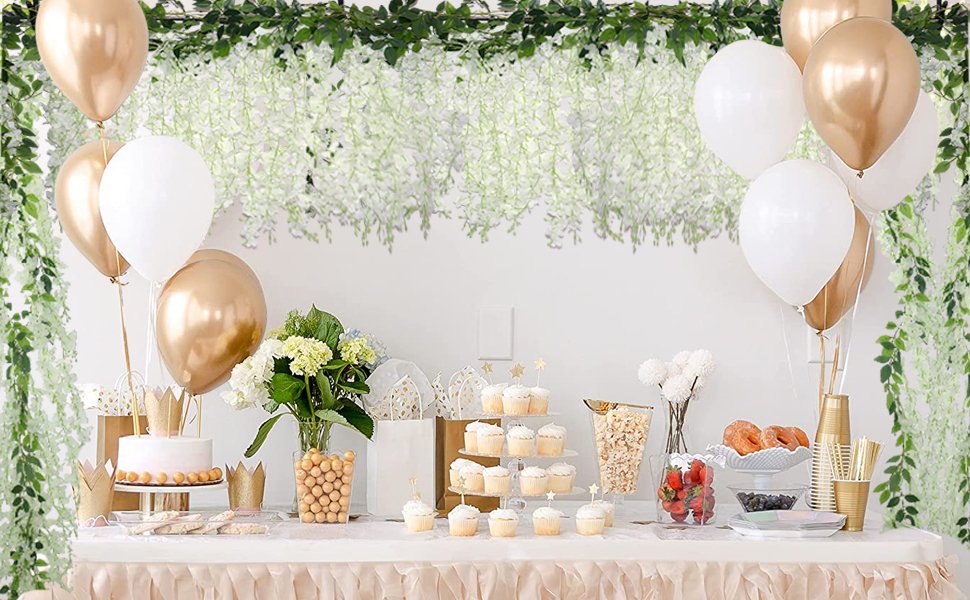 Party table with cupcakes, cake, donuts, and balloons, decorated with white wisteria and green garlands.