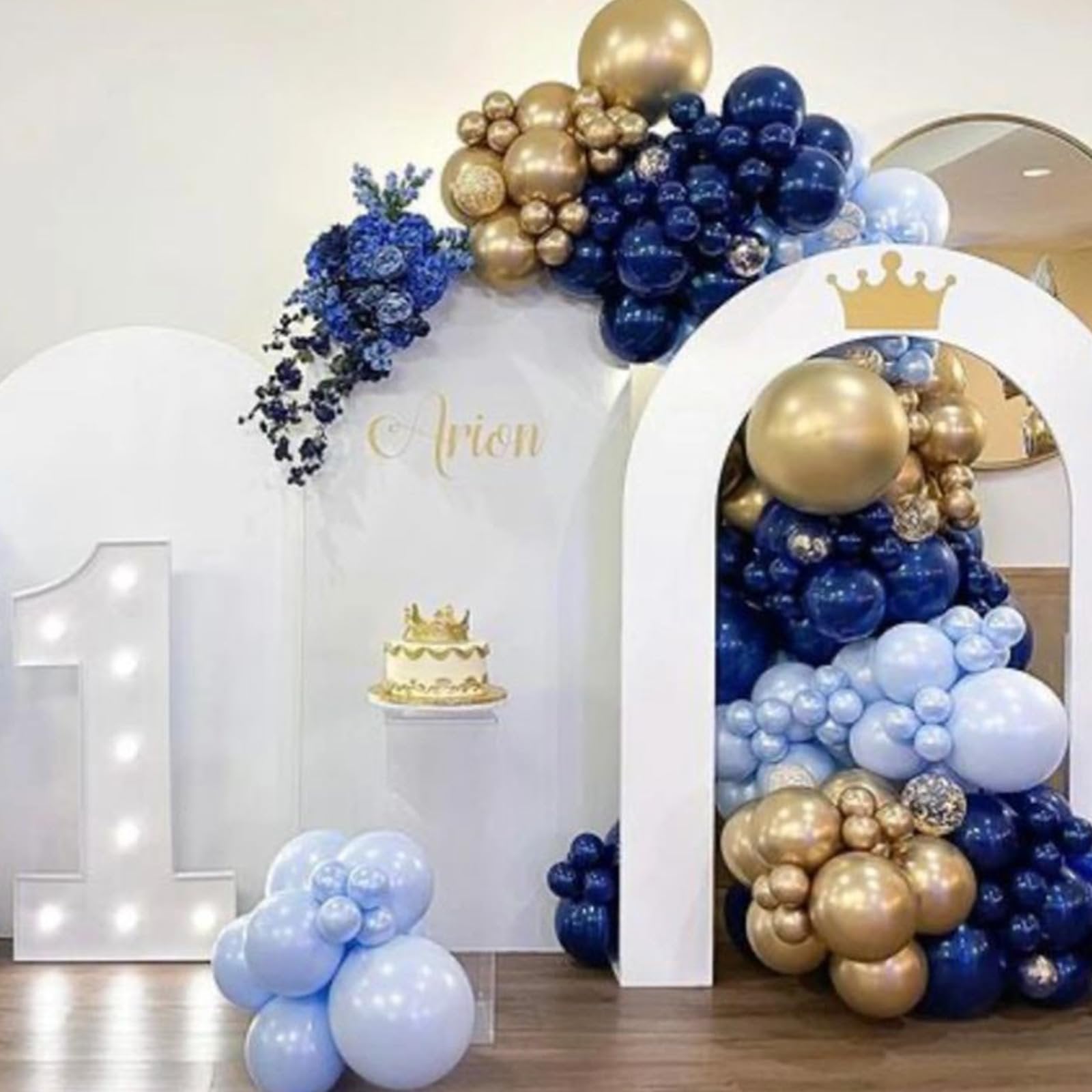 Birthday party decor with navy blue, gold, and light blue balloons, a cake with a crown, and a large illuminated number '1'.