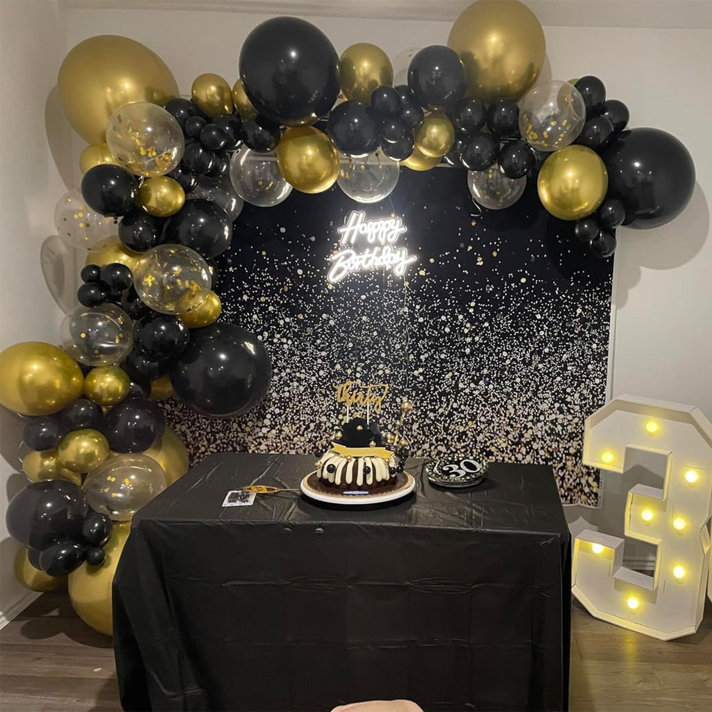 Birthday party setup with black and gold balloons, a cake, and a 'Happy Birthday' neon sign.