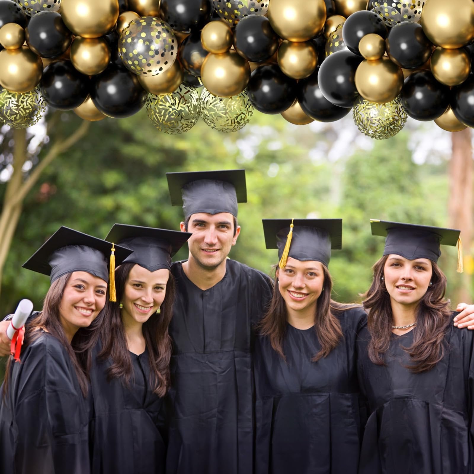 Five smiling graduates in black gowns and caps pose outdoors with a balloon arch overhead.