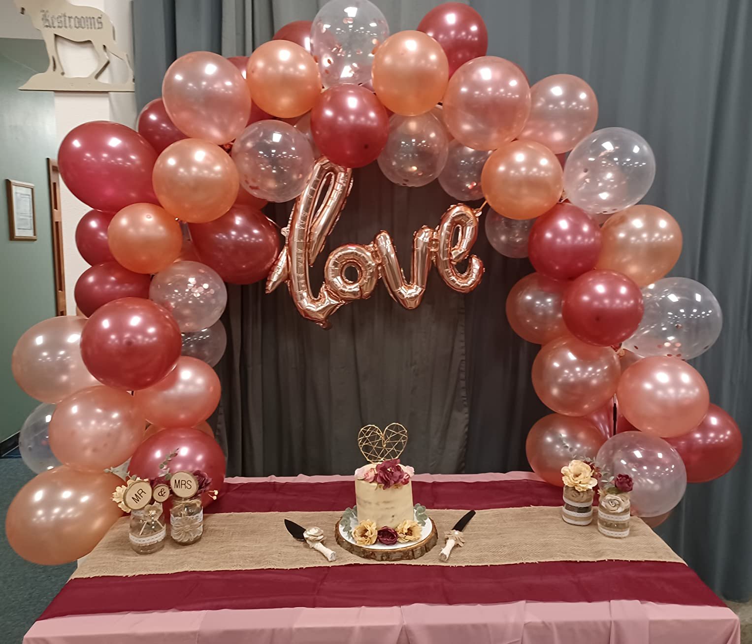 Balloon arch with 'love' foil balloon over a cake, with 'MR' and 'MRS' jars on a table.