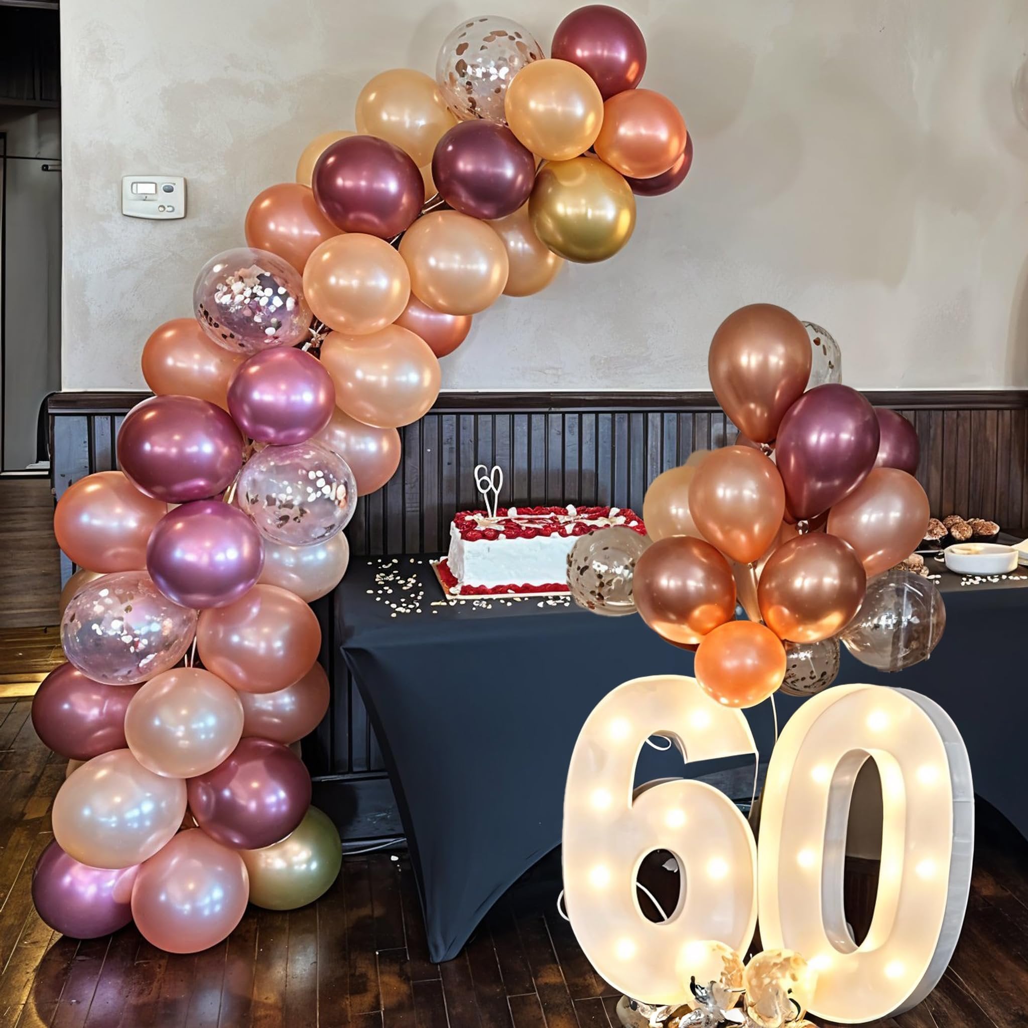 Party balloons in burgundy, rose gold, wine red, and champagne colors arranged in an arch and cluster, with a cake and illuminated number 60.
