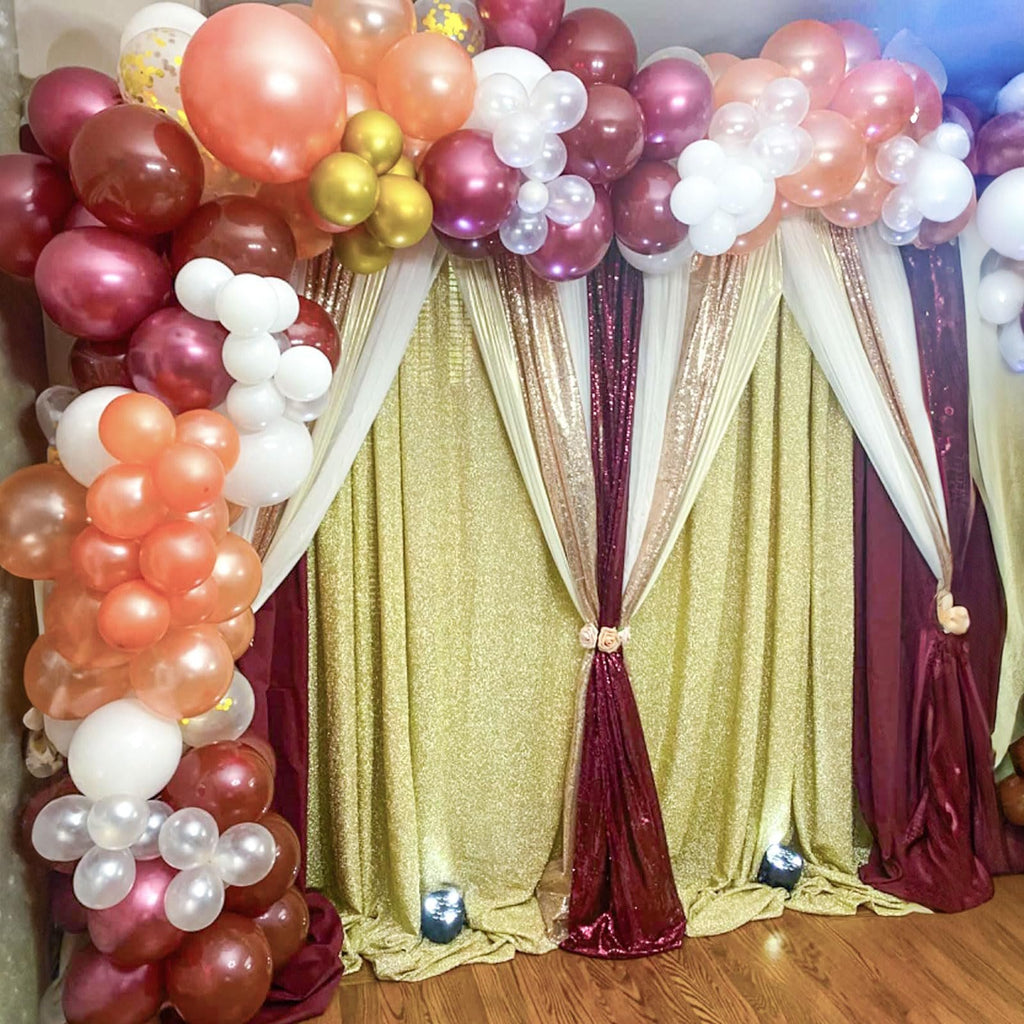 Balloon arch with burgundy, rose gold, wine red, and champagne latex balloons and confetti balloons, set against a backdrop of gold and burgundy sequined curtains.