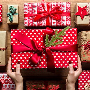 Hands holding a large red gift box with white hearts and a green pine sprig, surrounded by other wrapped gifts on a wooden surface.