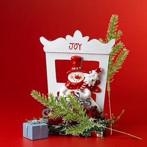 Christmas decoration with snowman figure, pine branches, gift box, and red accents on a red background.