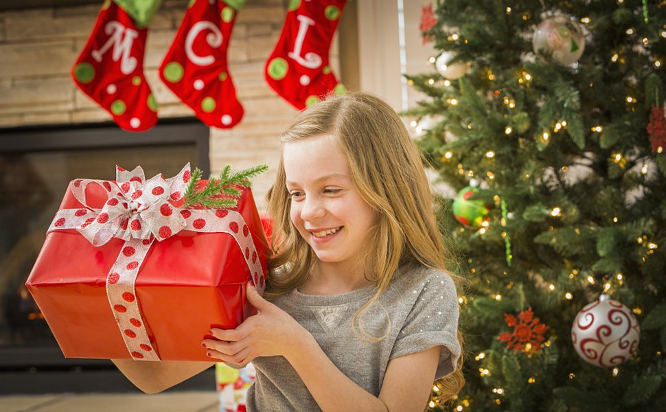 Smiling girl holding a red gift box with a polka dot ribbon in front of a Christmas tree and stockings