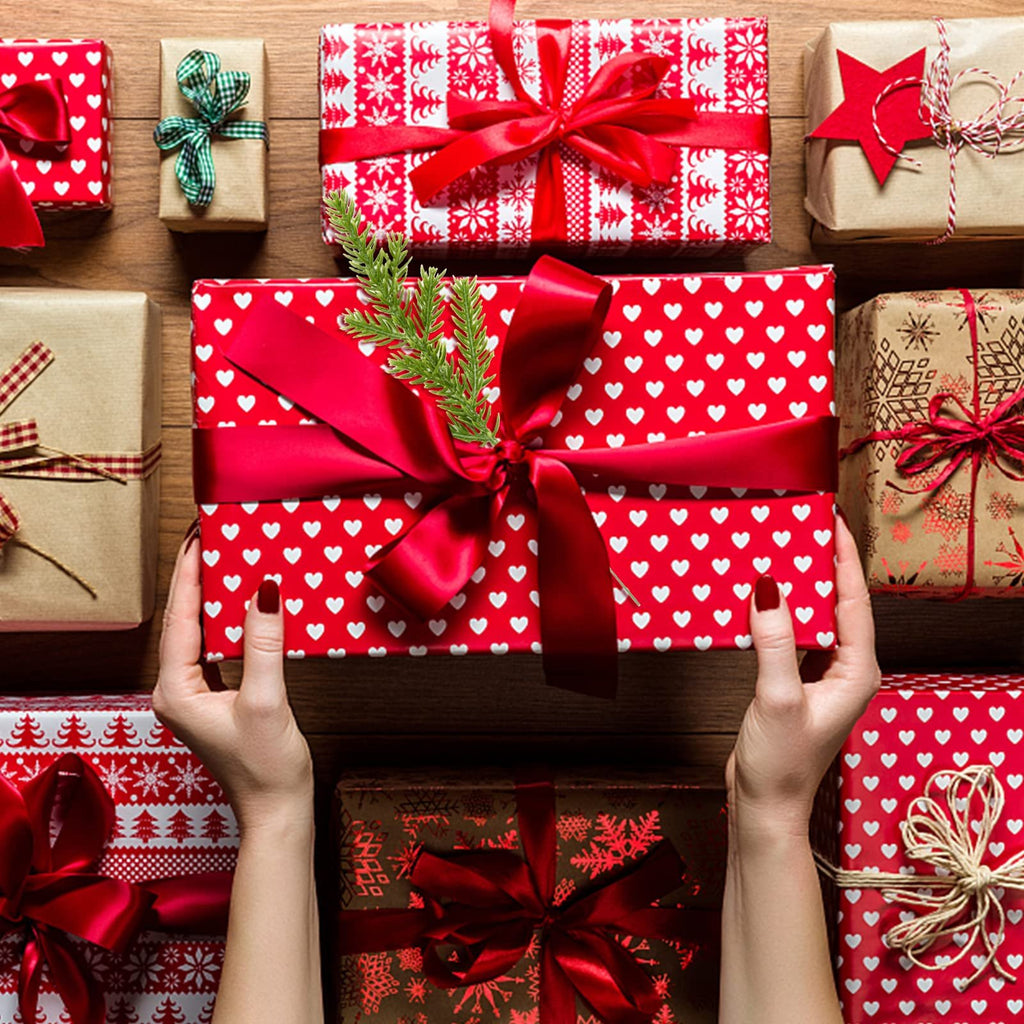 Hands holding a red gift box with white hearts, surrounded by other wrapped gifts and a sprig of pine needles.