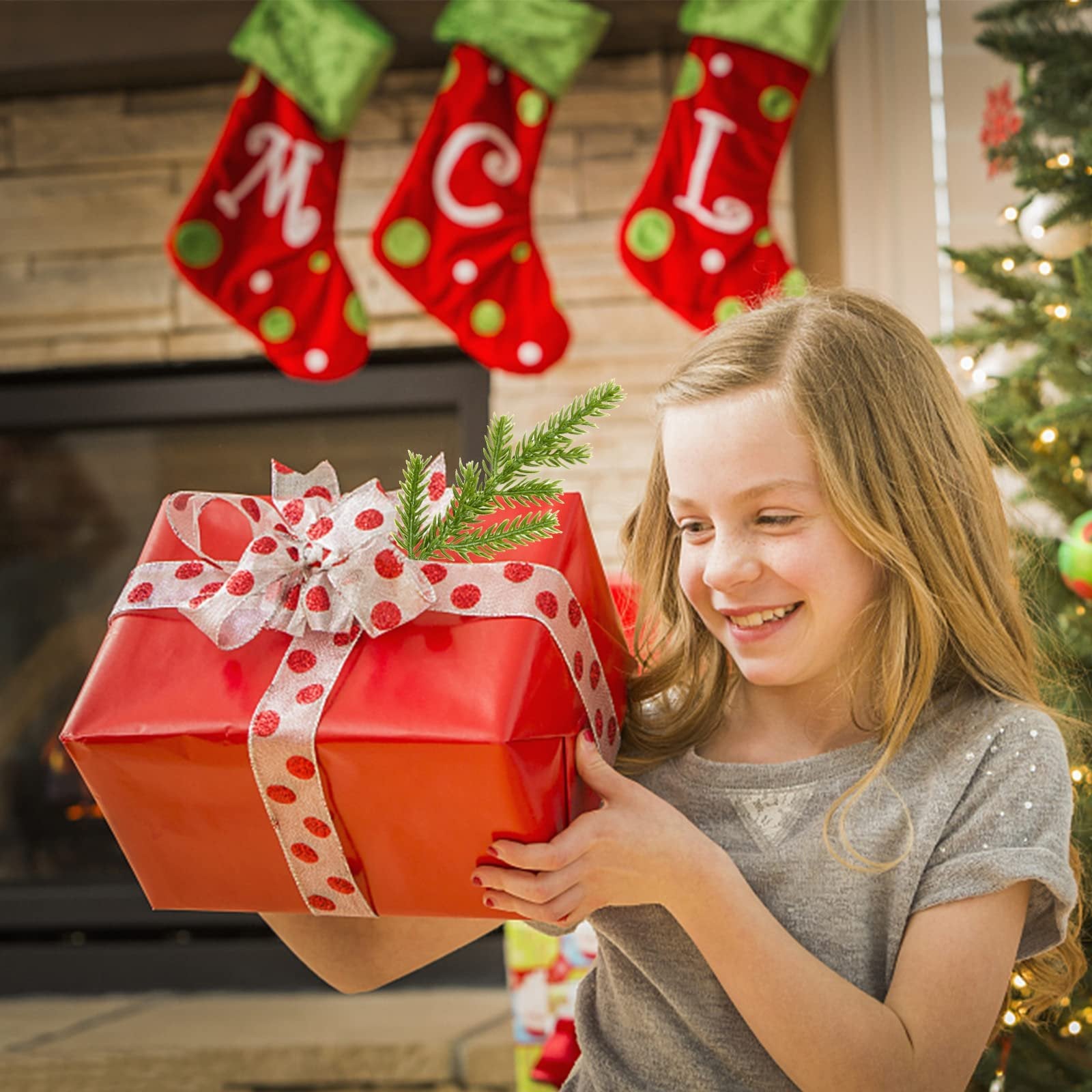 Young girl holding a red gift box with a festive ribbon and pine sprigs, with Christmas stockings and tree in background