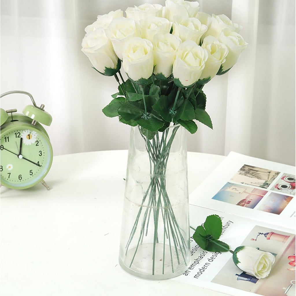Bunch of white artificial roses in a clear vase on a white table with a green alarm clock and an open magazine.