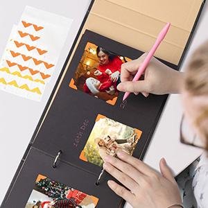 Person's hands using a pink pen to write in a black scrapbook photo album with photos and decorative stickers nearby.