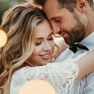 Close-up of a smiling bride in a white lace dress embracing a groom wearing a black bowtie, with soft bokeh lights.