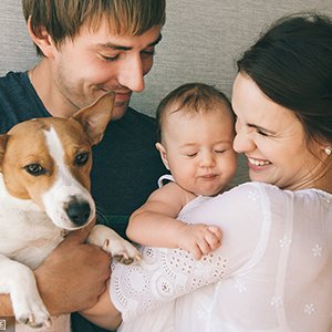 Family with baby and dog posing for a photo, smiling at each other.