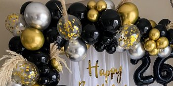 Party balloons in black, gold, and silver, some with confetti, arranged with dried grass and a 'Happy Birthday 26' banner.