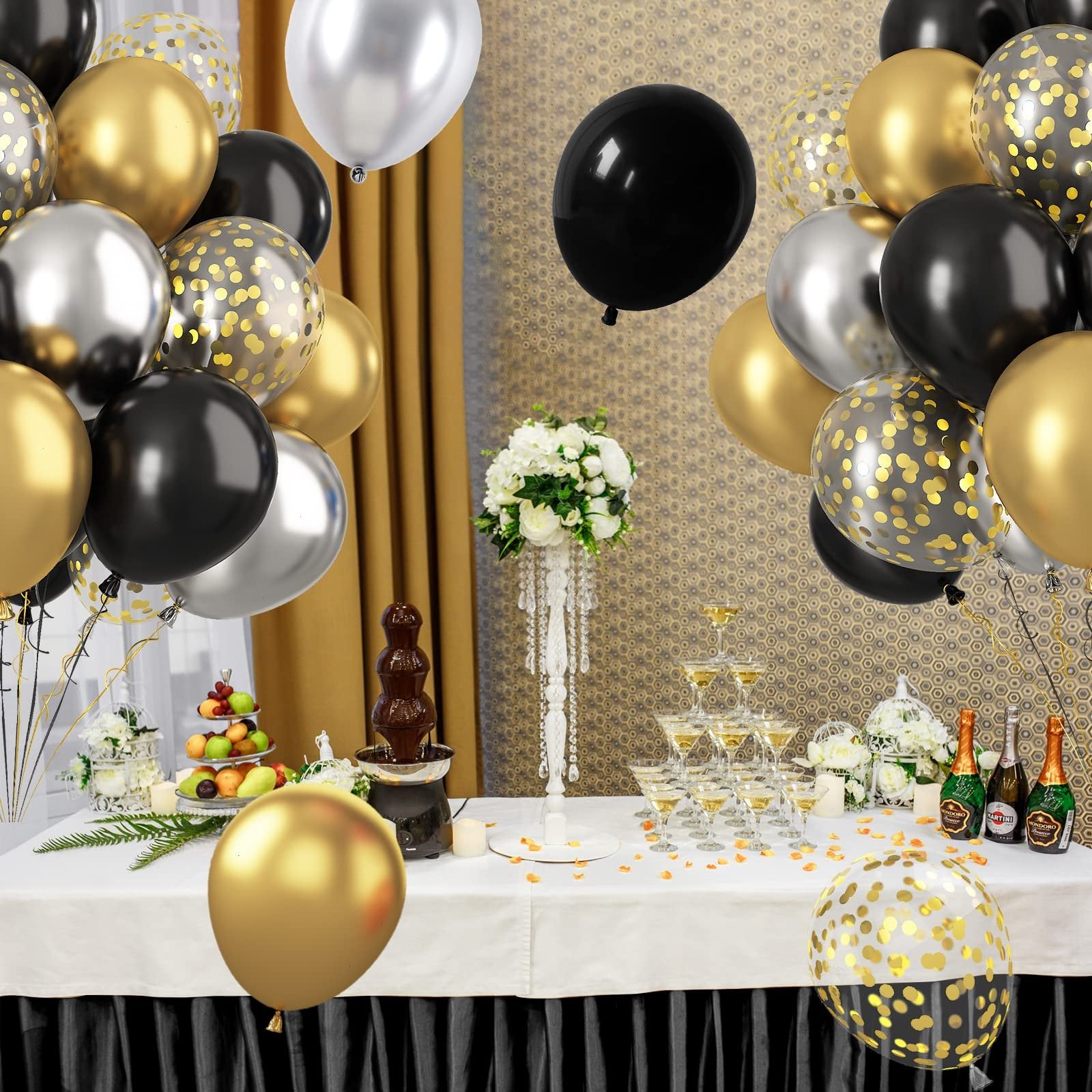 Party scene with black, gold, and silver balloons, champagne tower, chocolate fountain, and fruit display.