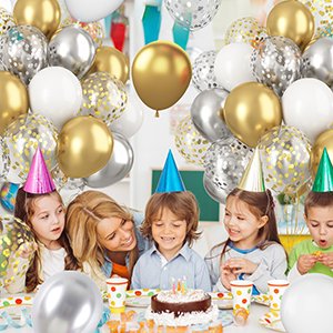 Group of children and an adult celebrating with metallic gold, silver, and white confetti balloons and a birthday cake.