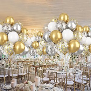 Metallic gold, silver, and white confetti balloons arranged in bouquets over a decorated event venue with tables and chairs.