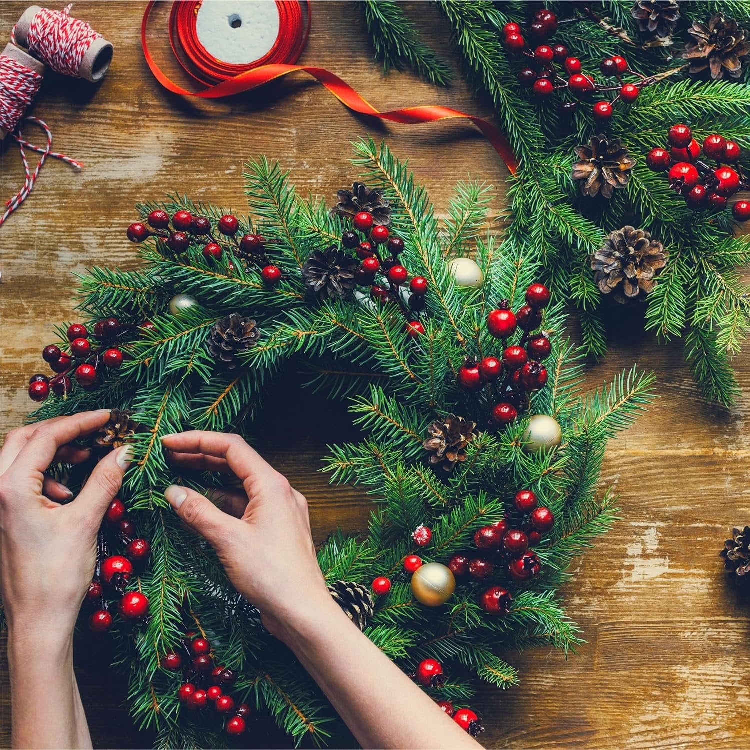 Hands assembling a Christmas wreath with pine branches, red berries, pinecones, and gold ornaments on a wooden table.