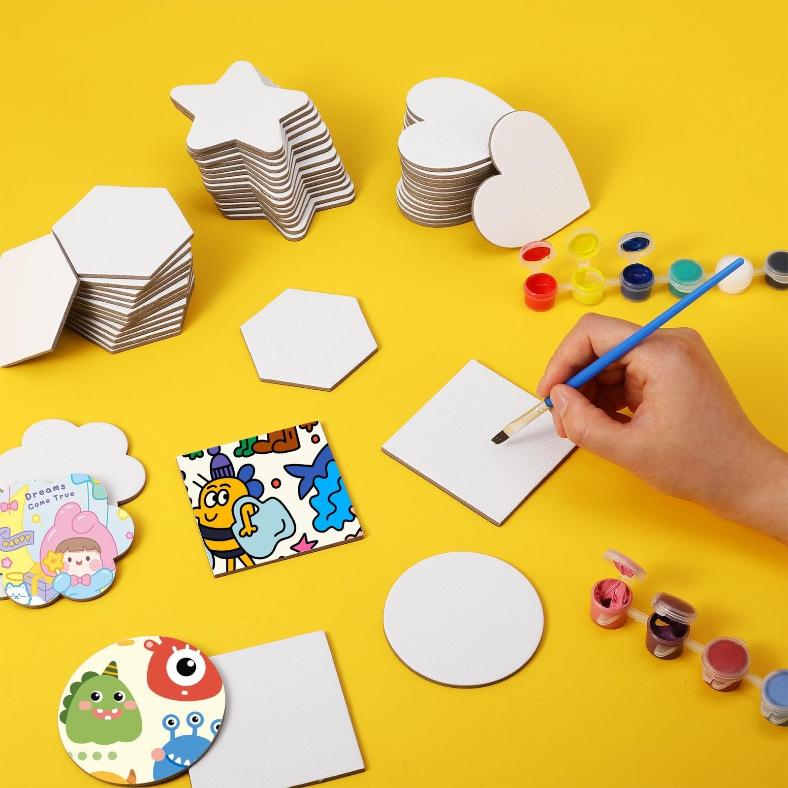 Hand painting a square canvas board with a brush, surrounded by stacks of blank canvas shapes and small paint pots.