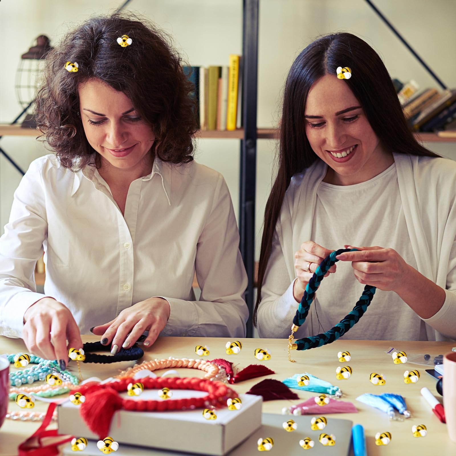 Two women crafting jewelry with tiny resin bees scattered around them and on their hair.