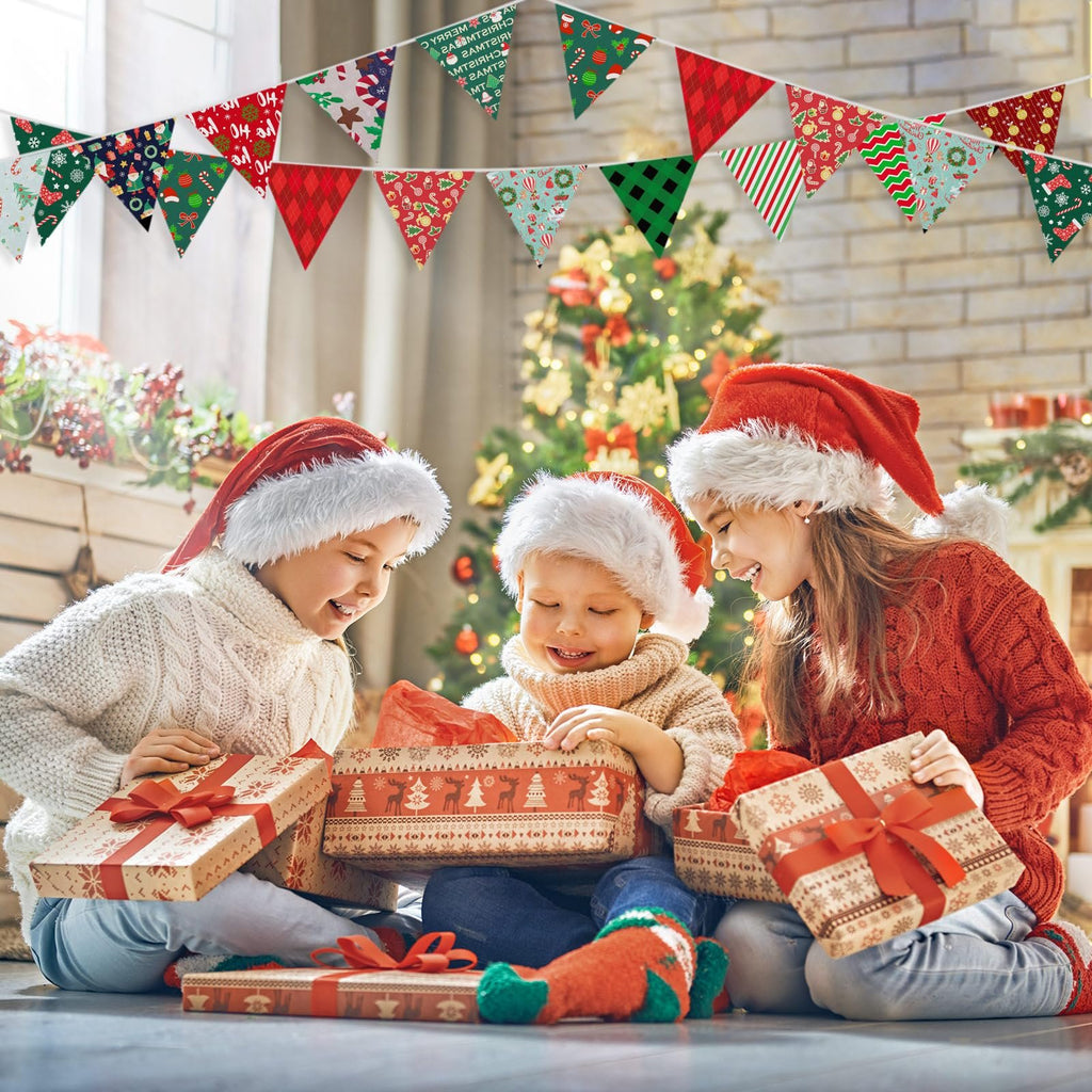 Three children in Santa hats opening Christmas gifts with a decorated tree in the background and festive bunting overhead.