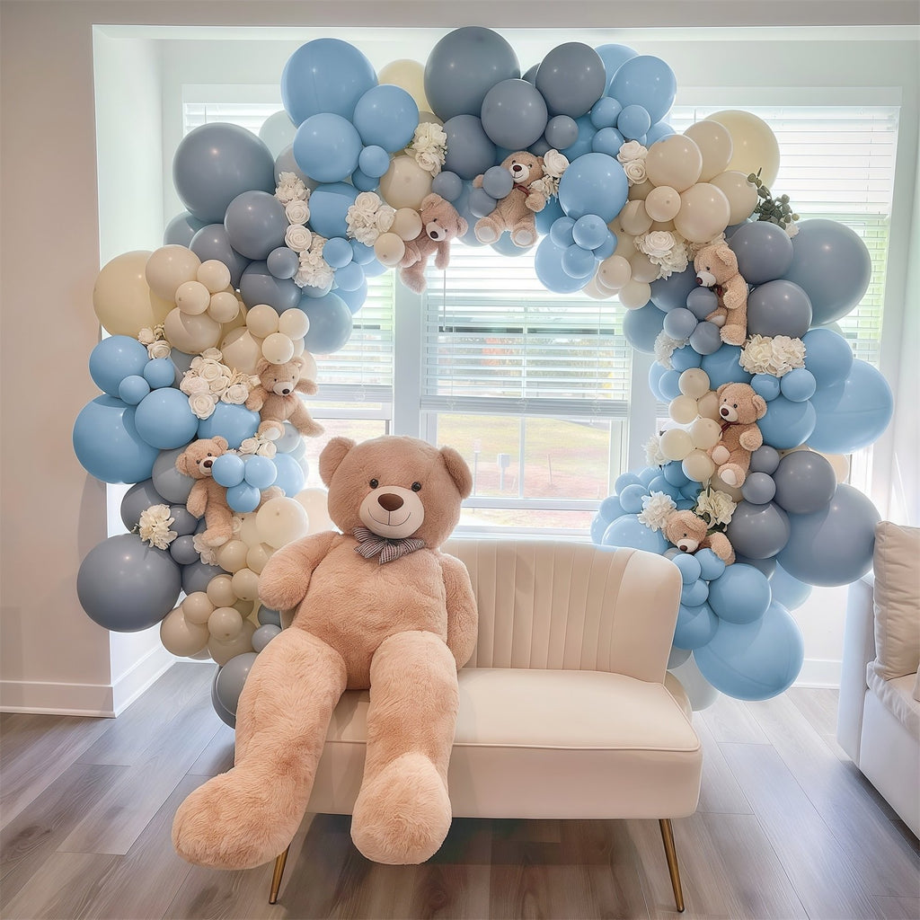 Dusty blue, white, and champagne gold balloon arch with teddy bears and white flowers, framing a large teddy bear on a cream sofa.