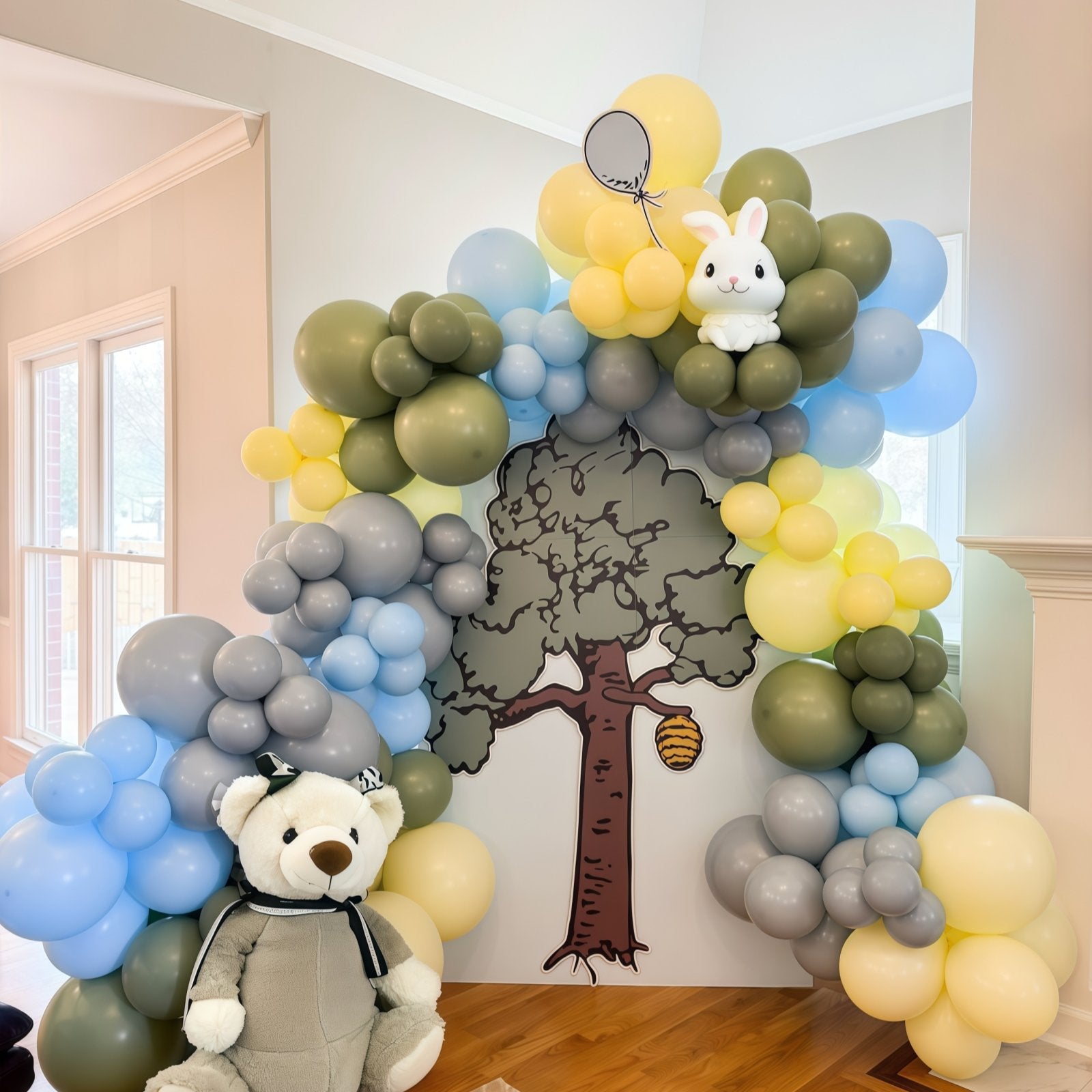 Balloon arch with a tree cutout and a teddy bear in front, featuring dusty blue, olive green, yellow, and gray balloons.