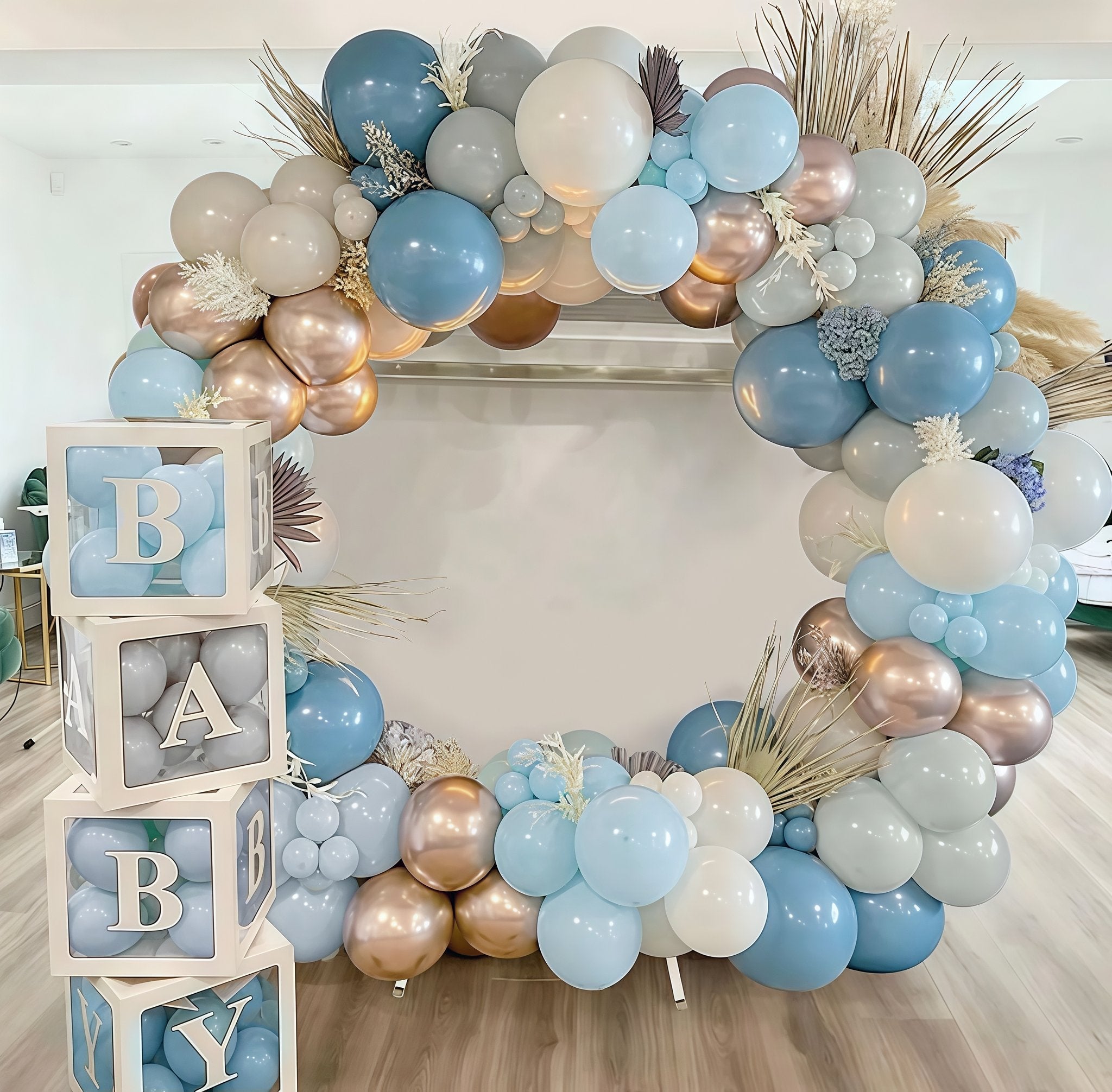 Dusty blue, sand, and champagne gold balloon arch with decorative dried grasses and white snowflake accents, next to stacked "BABY" blocks.