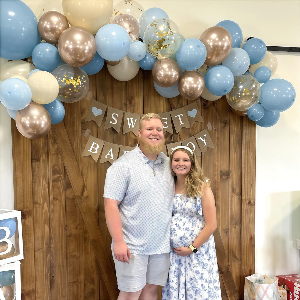 Couple posing in front of a wooden wall decorated with a balloon arch and 'SWEET BABY BOY' banner
