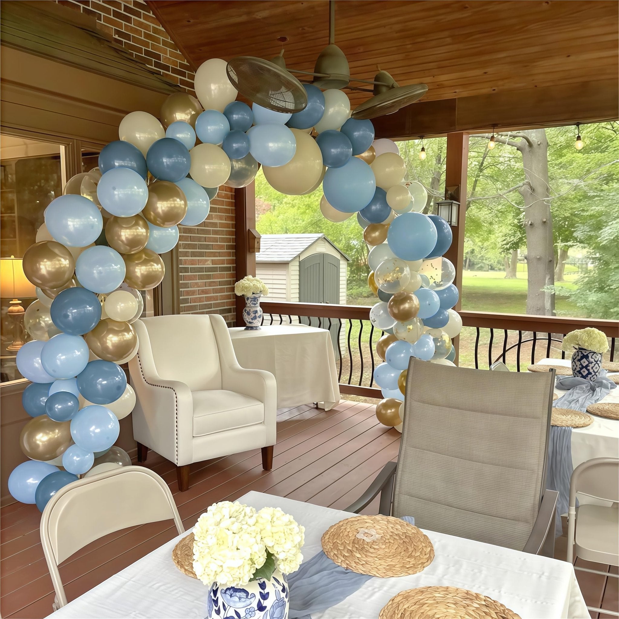 Balloon arch in dusty blue, white, and gold over a decorated outdoor patio with tables and chairs