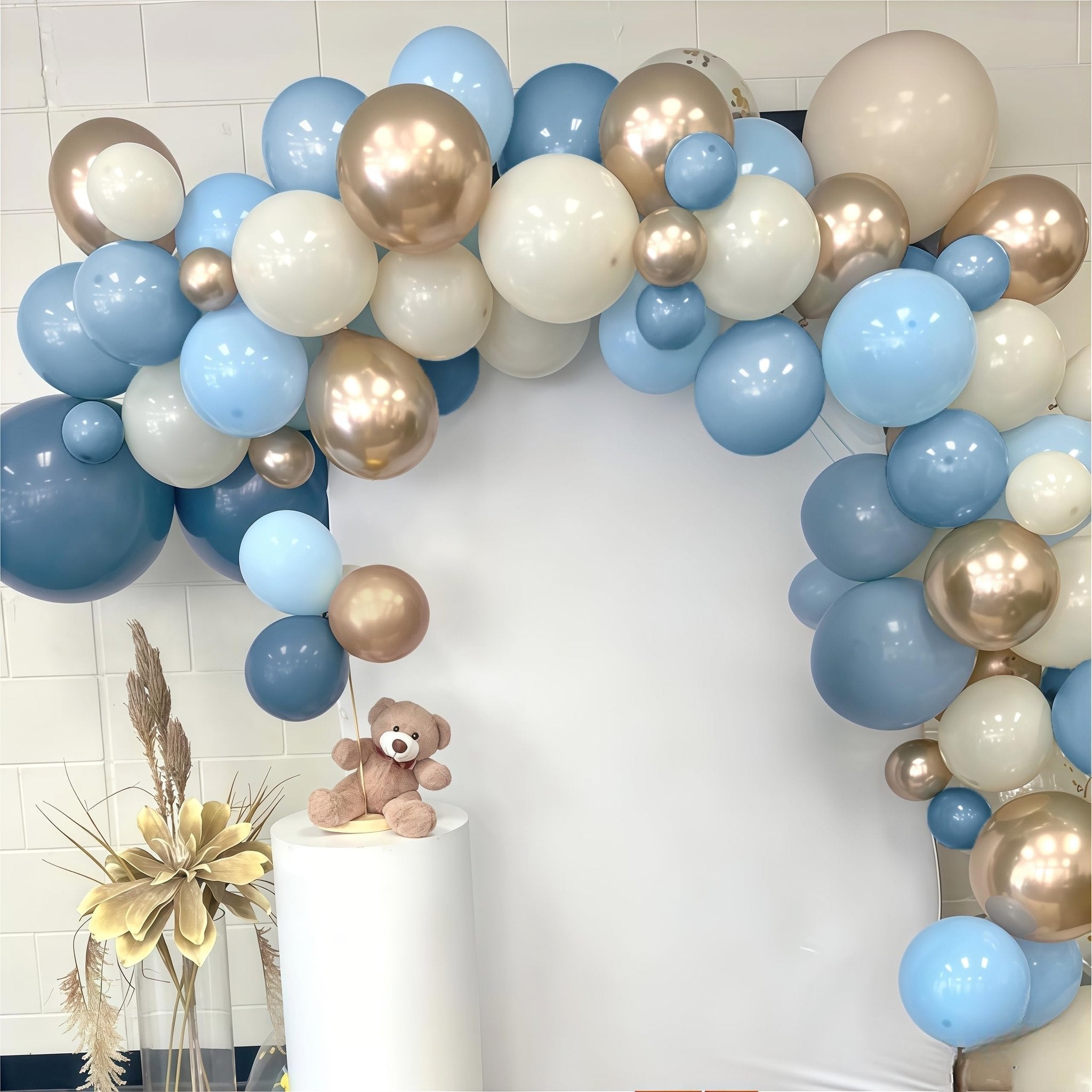 Dusty blue, white, and champagne gold balloon arch with a teddy bear on a pedestal and decorative reeds.