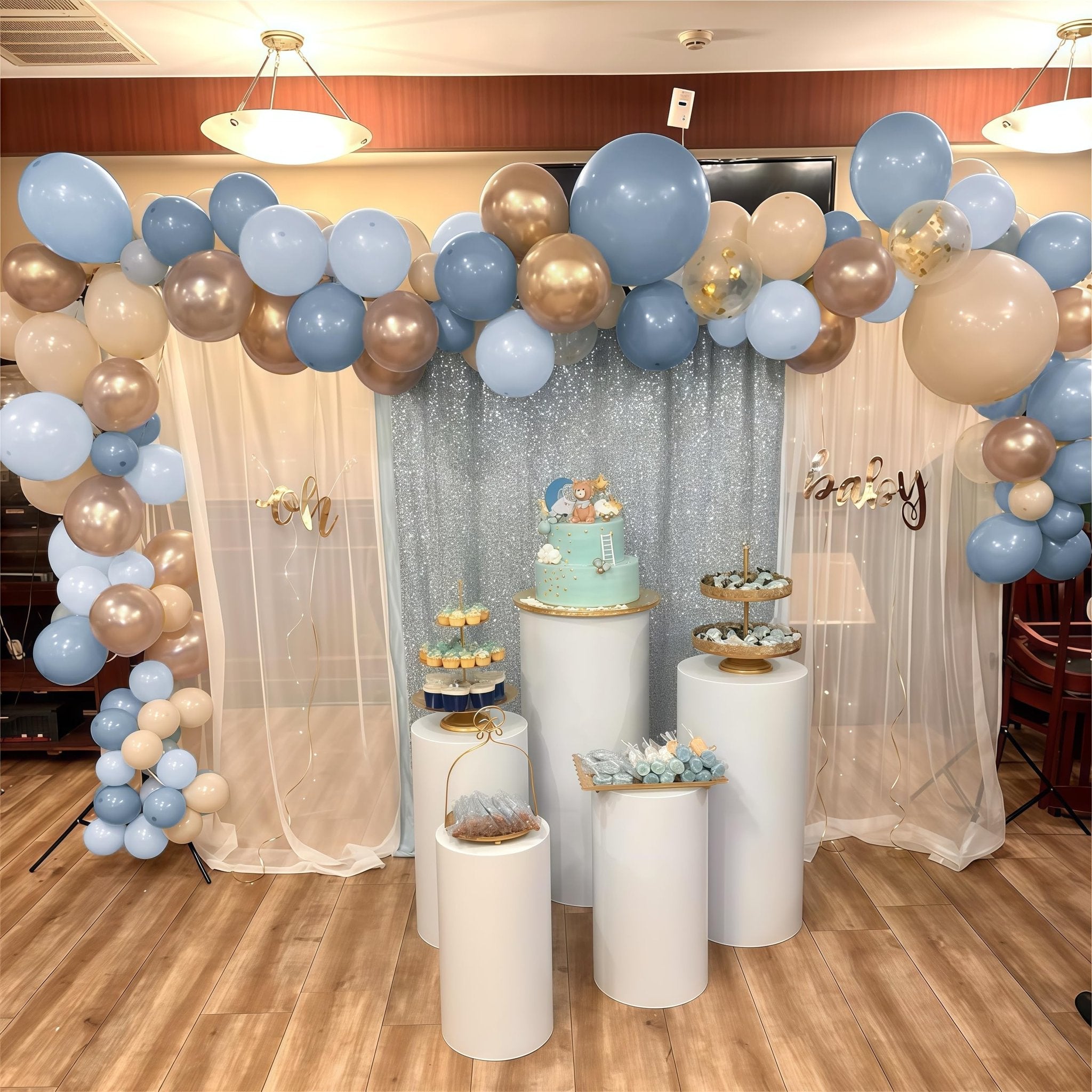 Dusty blue, champagne gold, and white balloons arching over a cake and cupcakes at a baby shower.