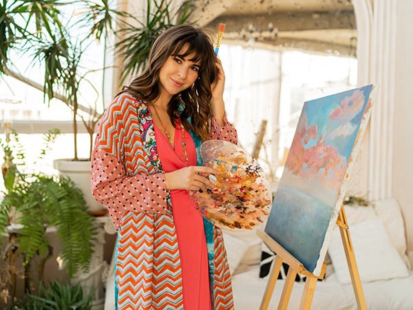 Woman holding a paint palette and brush, standing next to an easel with a painting.