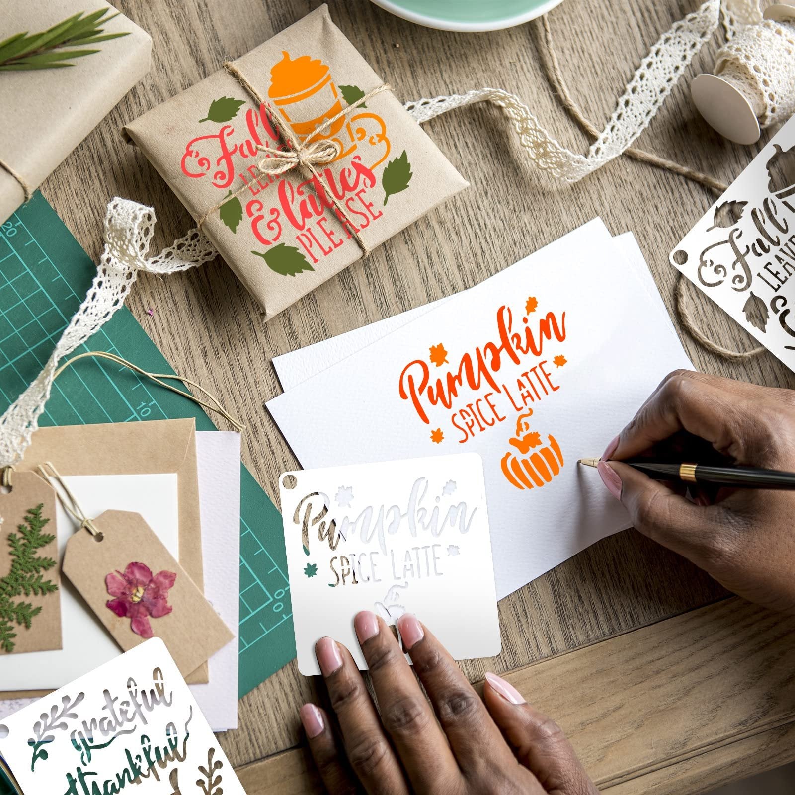 Hands using fall-themed stencils to decorate a gift-wrapped box and paper with pumpkin and leaf designs.