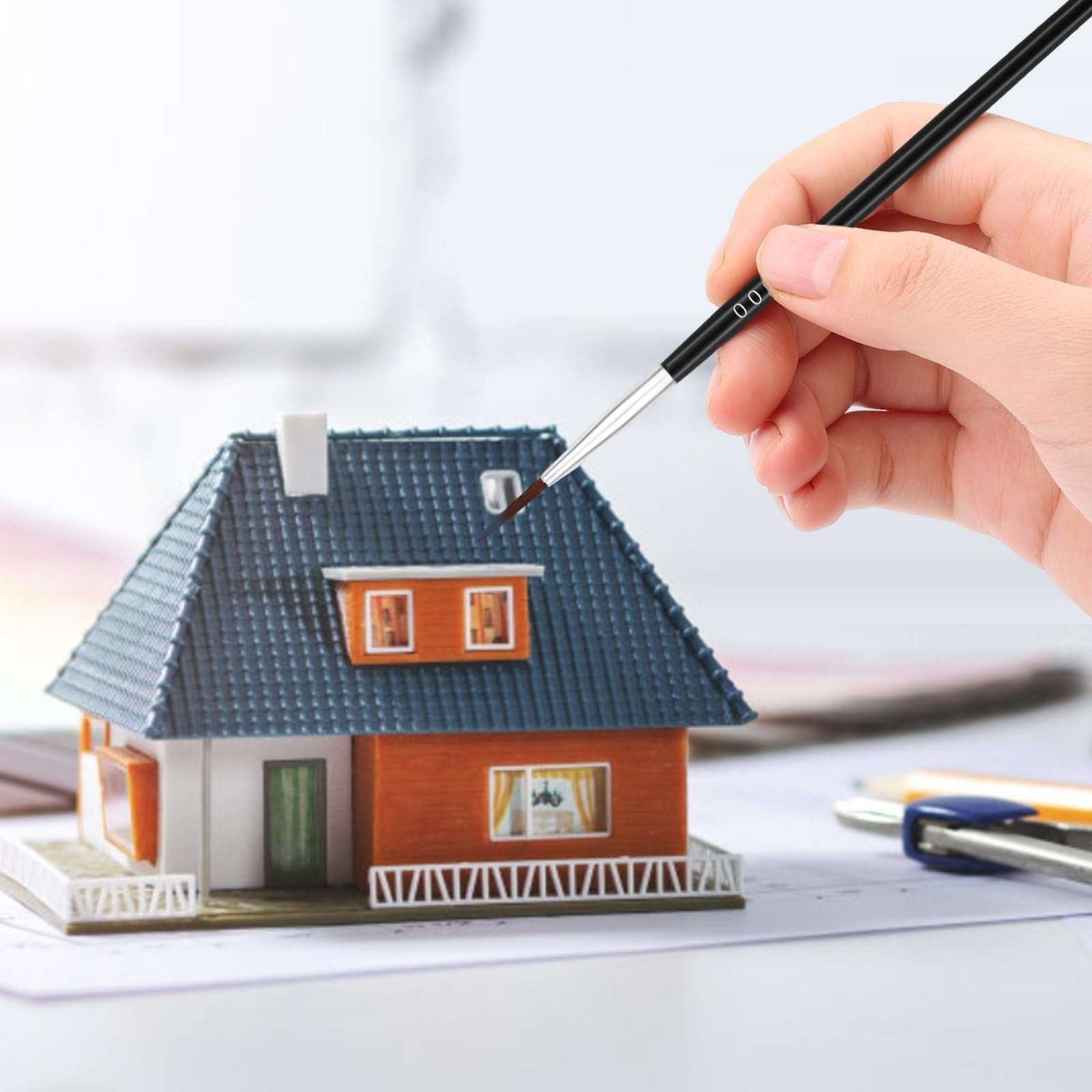 Hand holding a fine tip paint brush with size 00 marking, painting a miniature house model.