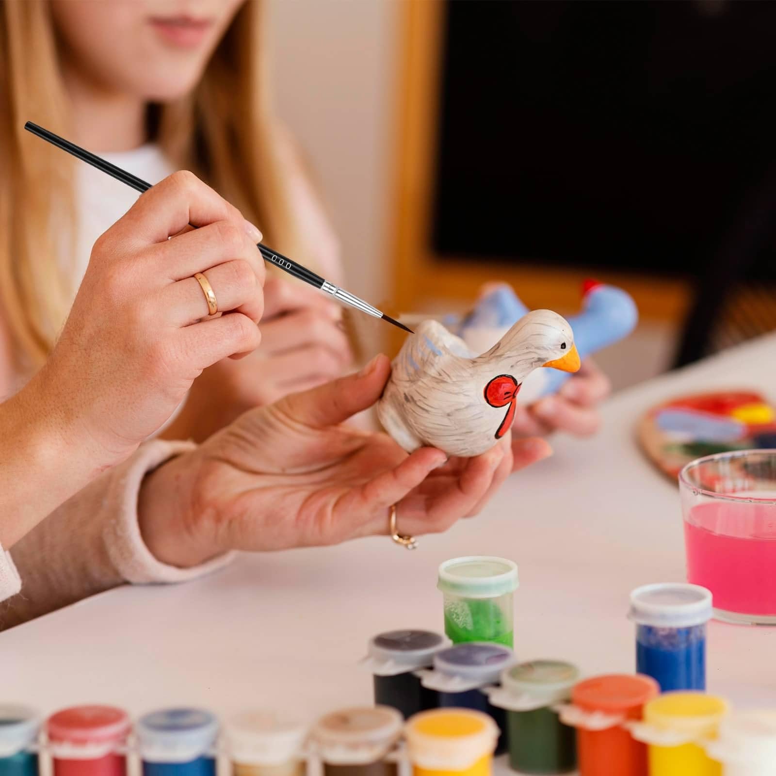 Close-up of hands painting a ceramic bird figurine with a fine-tip brush, surrounded by small paint pots and a glass of pink liquid.