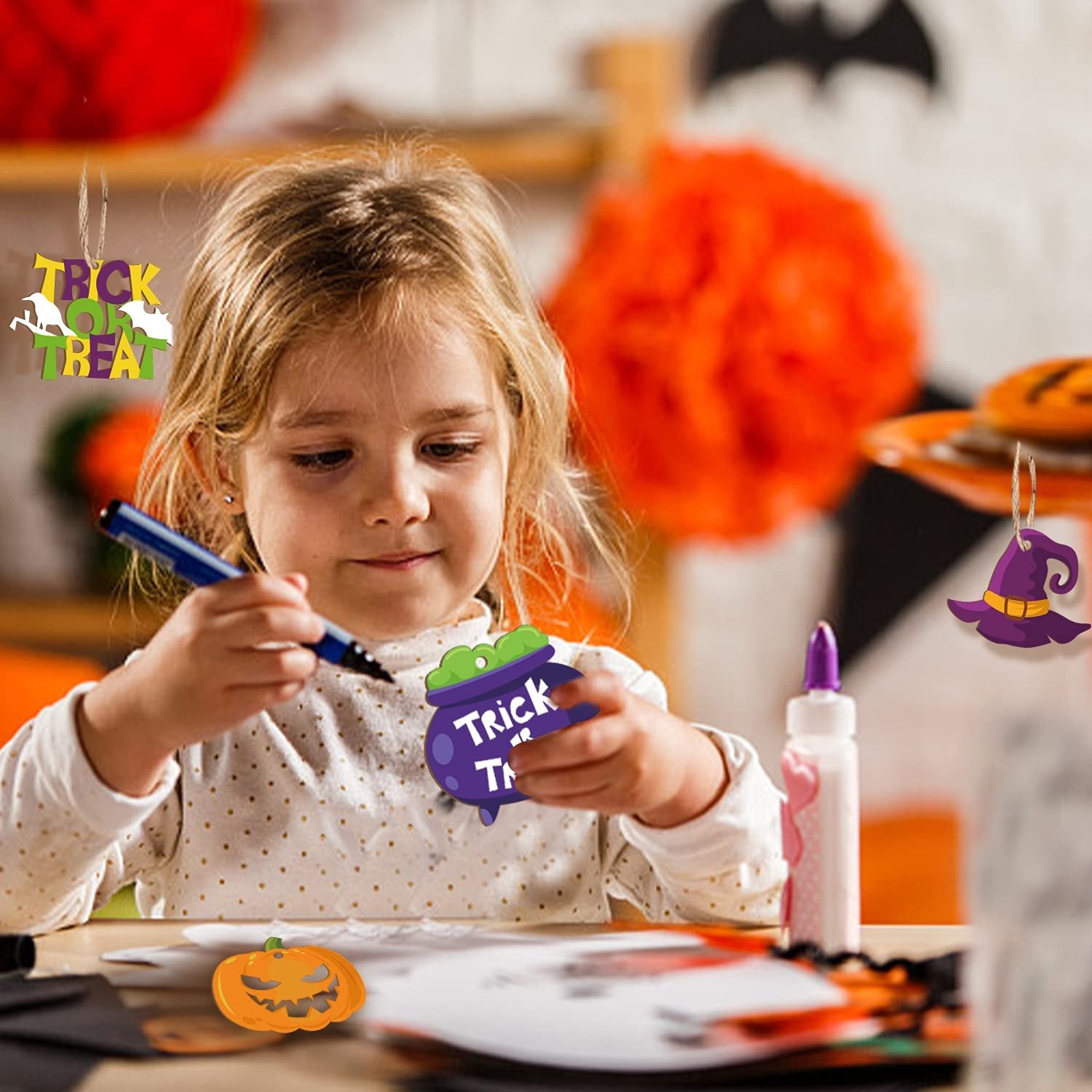 Young girl crafting Halloween ornaments, holding a purple cauldron ornament with 'Trick or Treat' text.