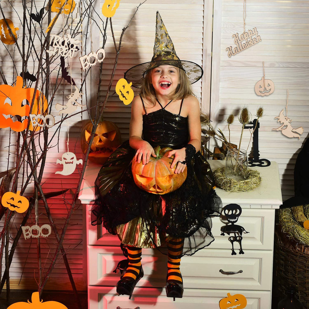 Smiling girl in witch costume holding a carved pumpkin, surrounded by Halloween decorations including wooden ornaments and paper cutouts.