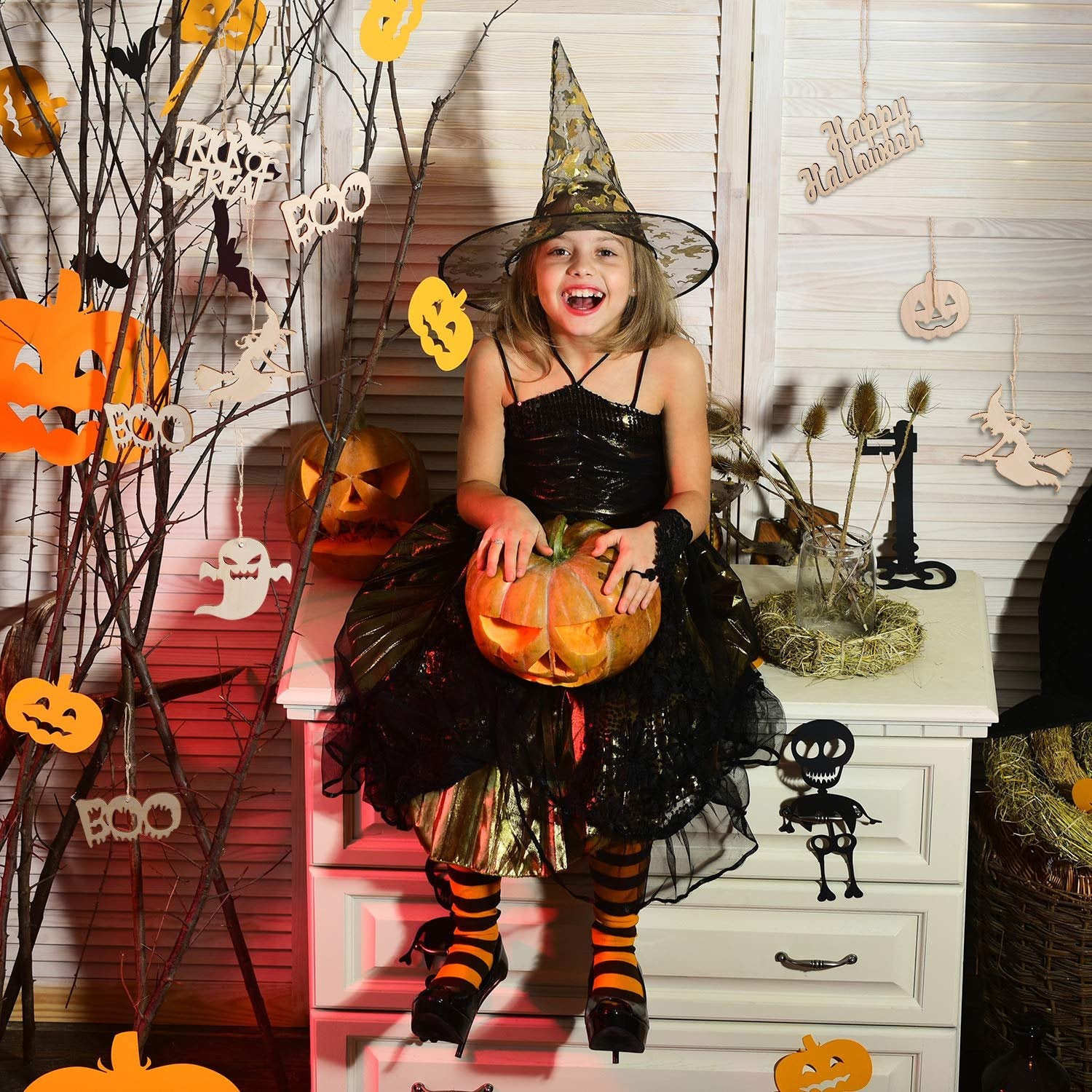 Smiling girl in witch costume holding a carved pumpkin, surrounded by Halloween decorations including wooden ornaments and paper cutouts.