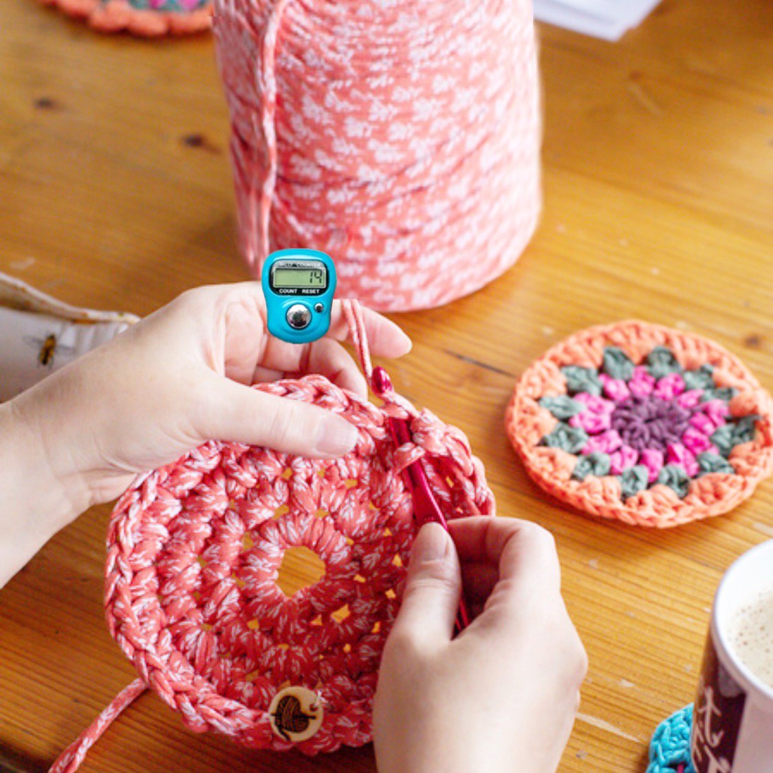 Hands crocheting a pink circular project with a blue digital row counter on a finger, large spool of yarn in background.