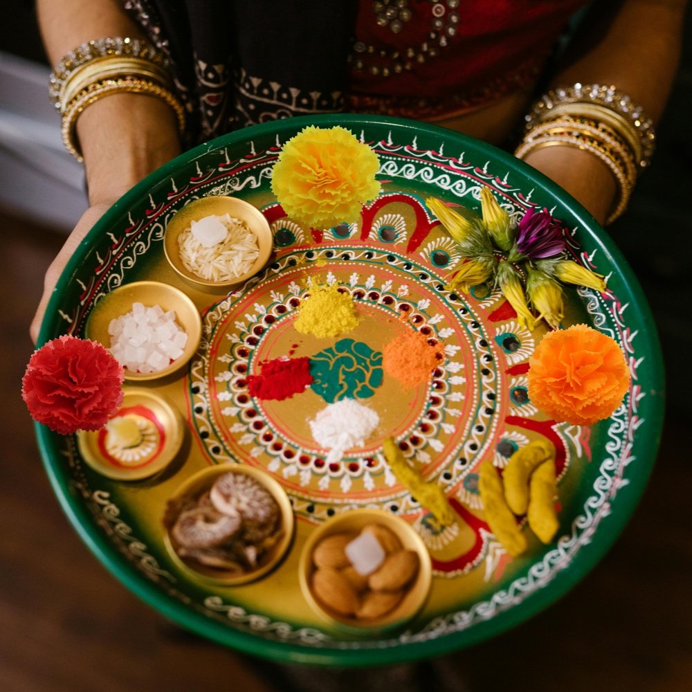 Person holding a decorative plate with colorful powders, rice, nuts, and marigold flowers for a cultural ceremony.