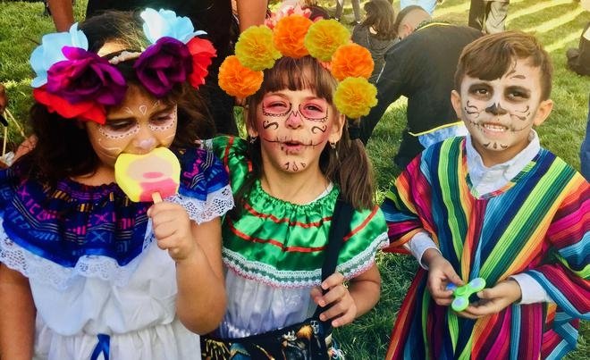 Three children in Day of the Dead face paint and festive attire, one eating a popsicle, one wearing a flower crown.