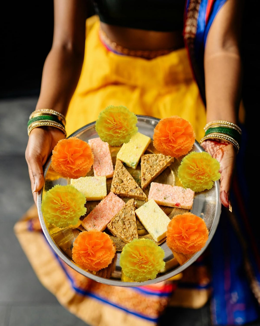 Person holding a plate of assorted sweets and artificial marigold flowers, decorated for a celebration.