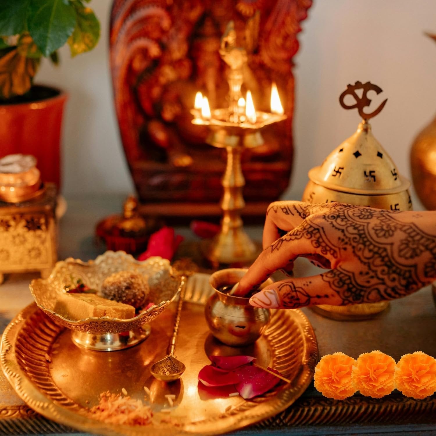 A hand with henna tattoos dips a finger into a small brass bowl on a decorative brass tray, with marigold flowers in the foreground.