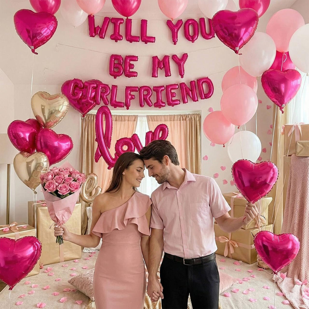 Couple in pink attire surrounded by heart and round balloons, rose petals, and gift boxes, with 'WILL YOU BE MY GIRLFRIEND' and 'LOVE' balloon text.