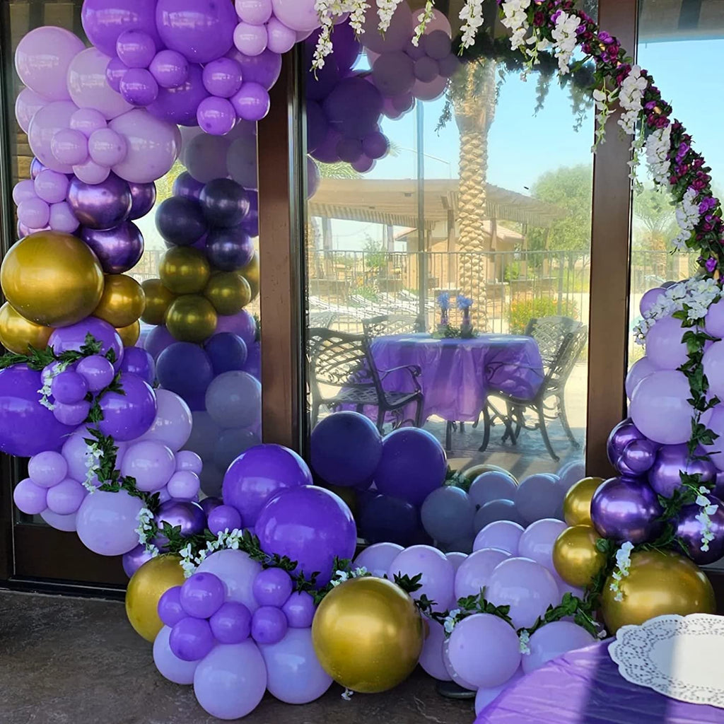 Purple and gold balloon arch and garland decoration with white flowers, set up outdoors near a patio table.
