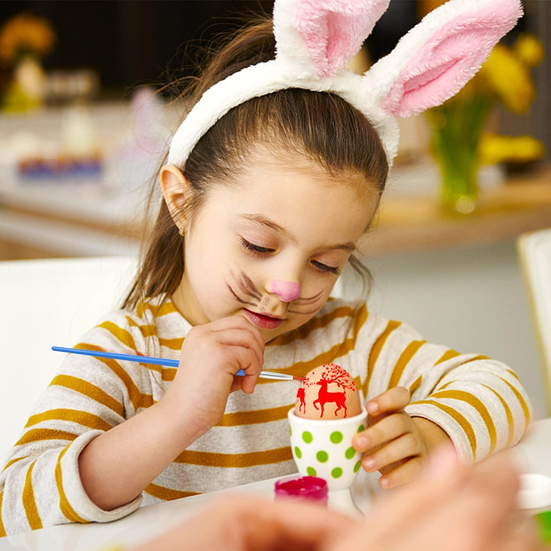Young girl with bunny ears painting an Easter egg with a red deer design