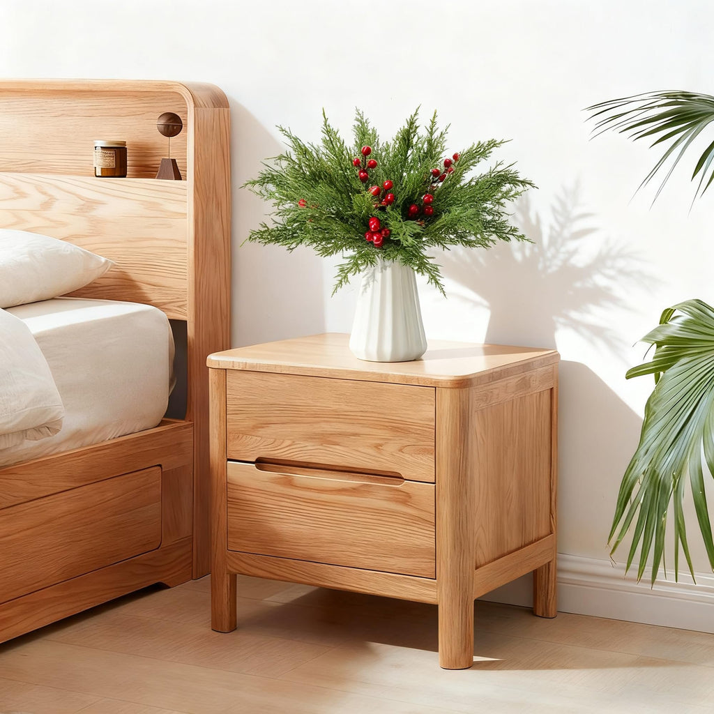 Artificial pine branches with red berries in a white vase on a wooden nightstand next to a bed.