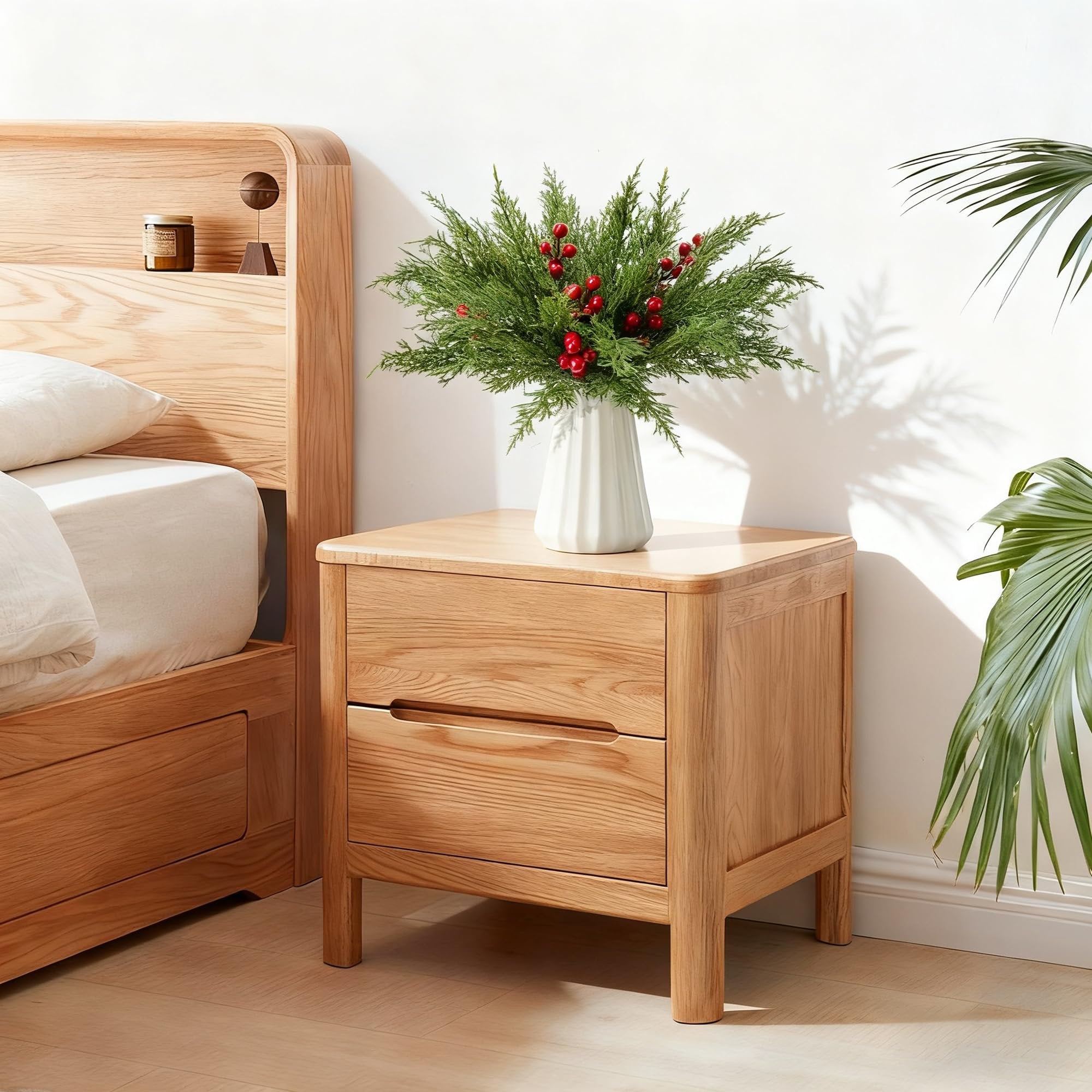 Artificial pine branches with red berries in a white vase on a wooden nightstand next to a bed.